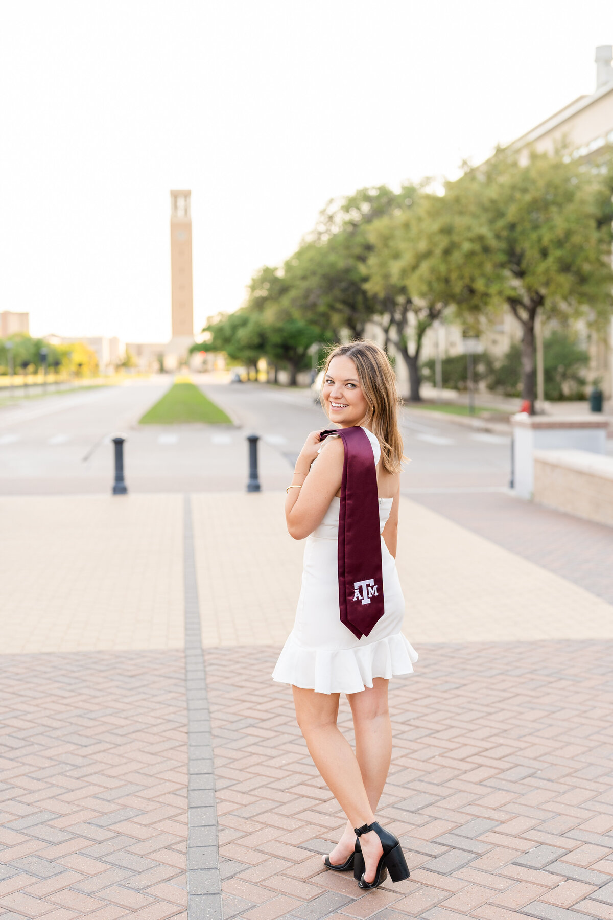 Texas A&M senior girl in white dress and maroon stole looking over shoulder with Bell Tower in the background