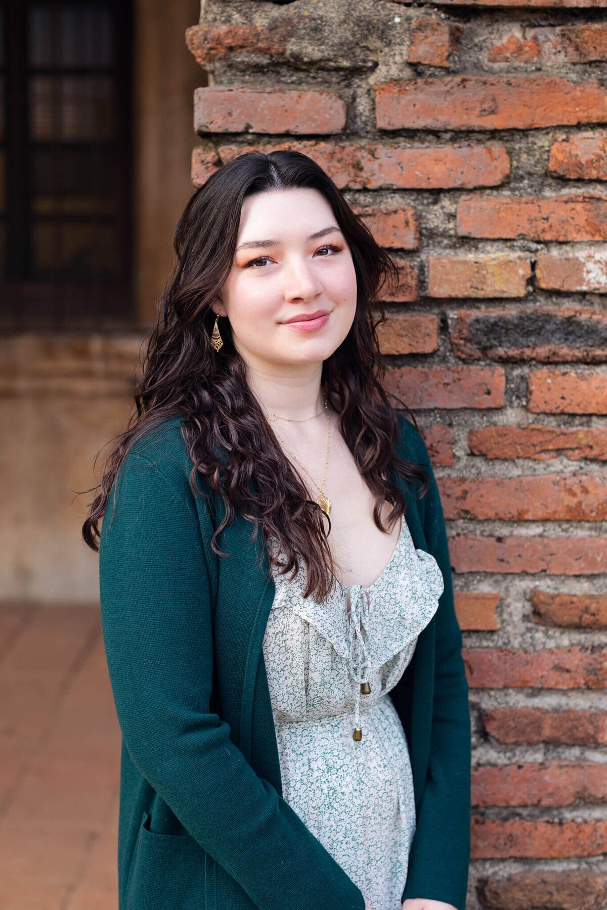 A young woman with long black hair leaning against a red brick wall, wearing a green cardigan and smiling softly with her lips at the camera.
