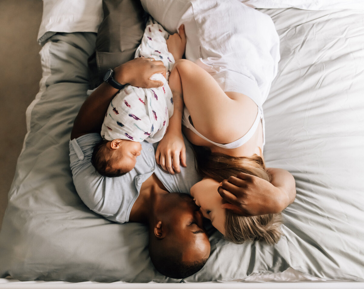 Newborn Photographer, Mom and dad laying on the bed and kissing while baby girl sleeps on dad's chest.