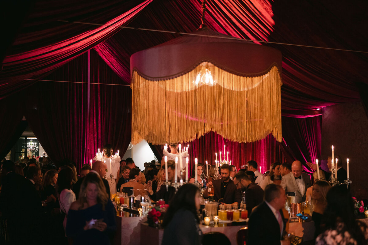 This stunning photo was captured at a tented wedding in Virginia by the talented photographer Elisabetta Marzetti, with Clayton Films providing videography services. In the image, two women are seen gracefully posing in black silk dresses, complemented by intricate black lace masks. The setting is a dimly lit, elegantly decorated room adorned with rich red drapes and ambient lighting, adding to the overall mysterious and sophisticated vibe of the scene.