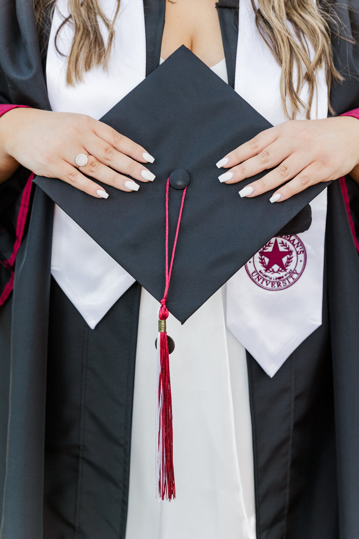 Close-up of a Texas State University graduate holding a black graduation cap with a maroon and white tassel. She wears a black gown with a white stole featuring the Texas State seal, and a white dress underneath. Her nails are painted white and she wears a round diamond ring.