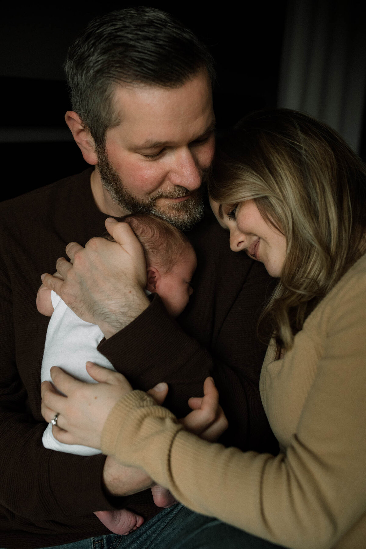 Dad hugging baby girl against his chest while mom gently leans into him while he softly looks at her.