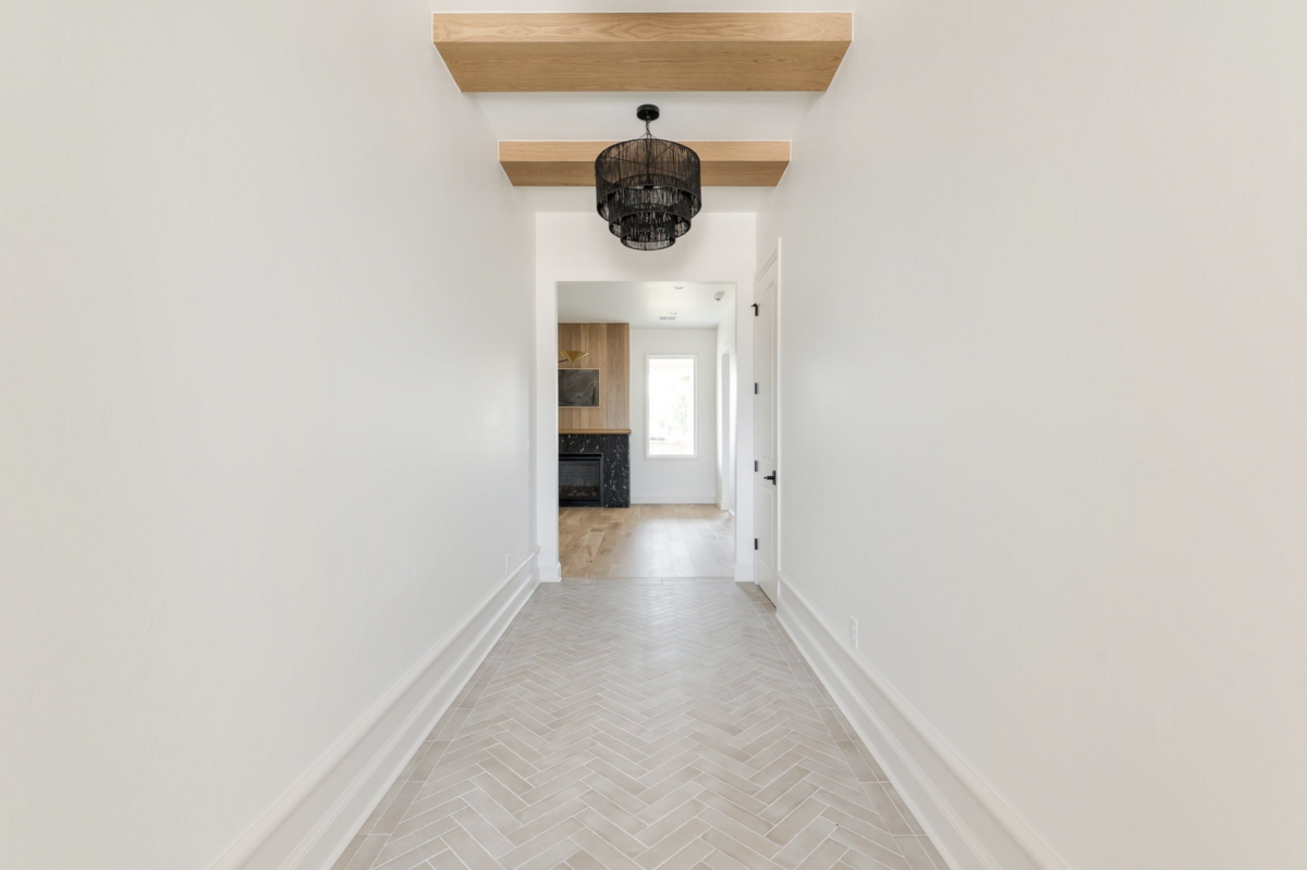 Bright entry hallway with light herringbone tile, white walls, natural wood ceiling beams, and a black tiered pendant leading into a living area with wood and marble fireplace surround.
