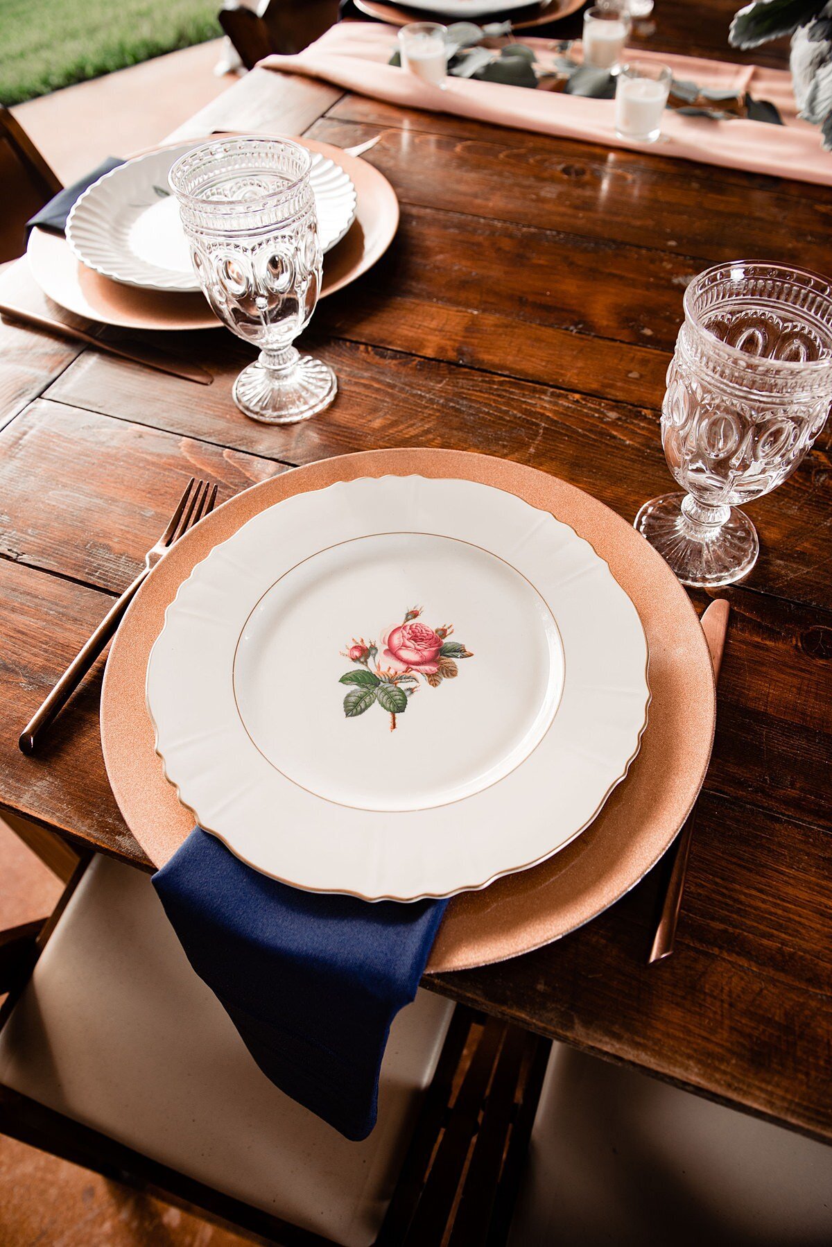 A brown barn wood farm table with a gold charger and vintage china plate is accented with a navy linen napkin and gold flatware with a clear glass water goblet.