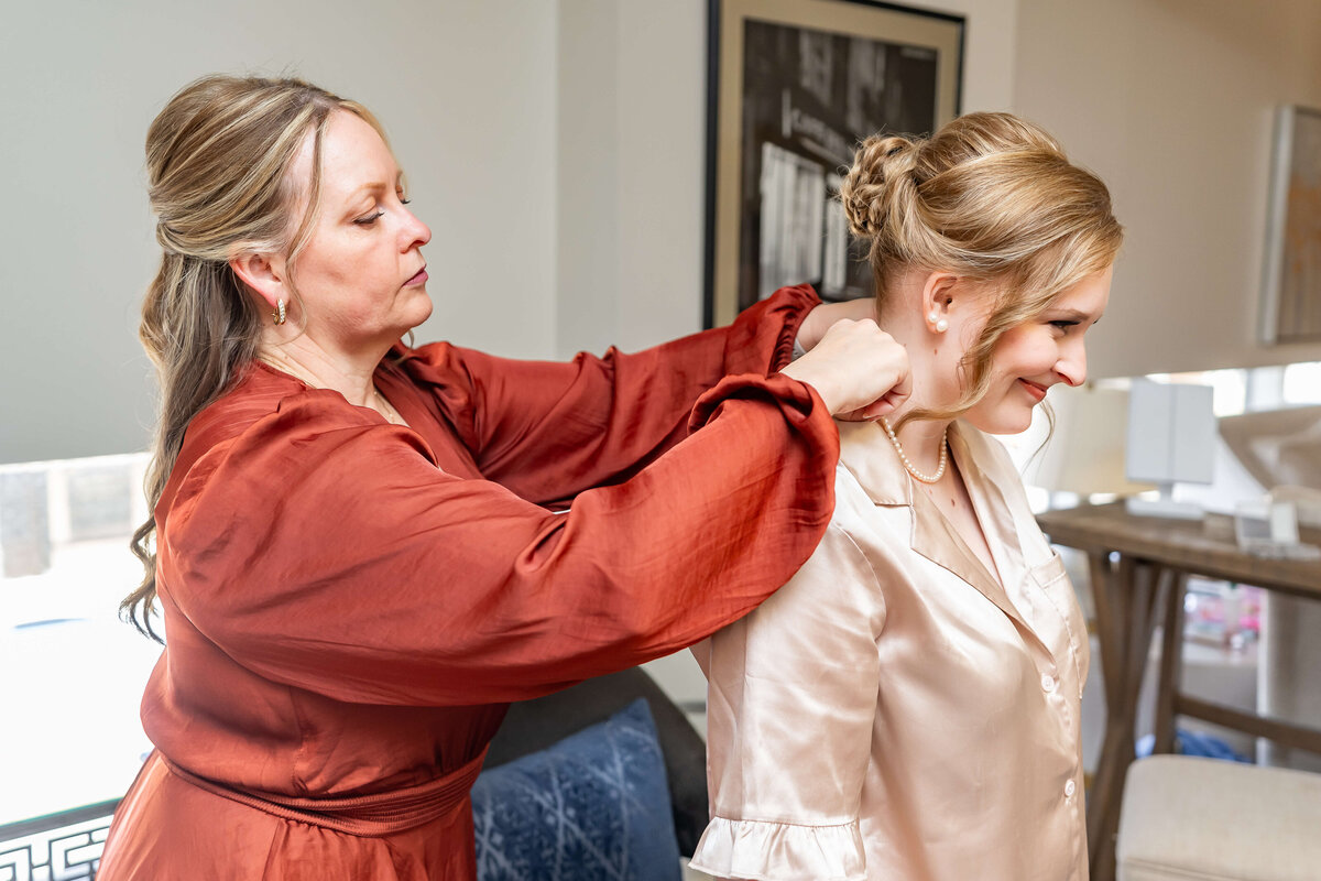 A woman in a rust-colored dress helps another woman in a cream satin outfit put on a necklace. They are indoors, and the woman receiving help is smiling.