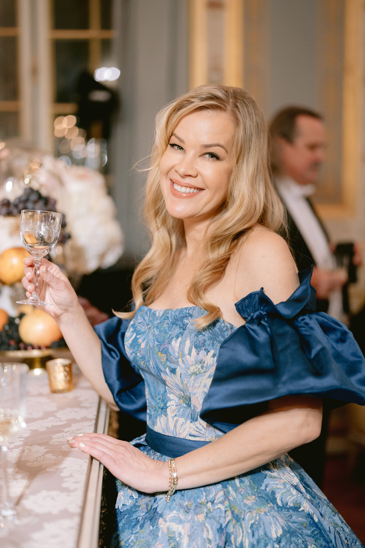 The photo was taken at Shangri La Hotel & Ritz Paris Hotel in Paris, France, by photographer Matteo Coltro. In the image, a woman with blonde hair smiles as she holds a glass of wine in her left hand. She is elegantly dressed in a blue off-the-shoulder dress adorned with floral patterns. Additionally, a man in the background, dressed in a suit and bow tie, is slightly blurred. The overall setting suggests a formal event or celebration, adding a touch of sophistication to the scene captured in the photograph. Videographers: Zen Film Works.