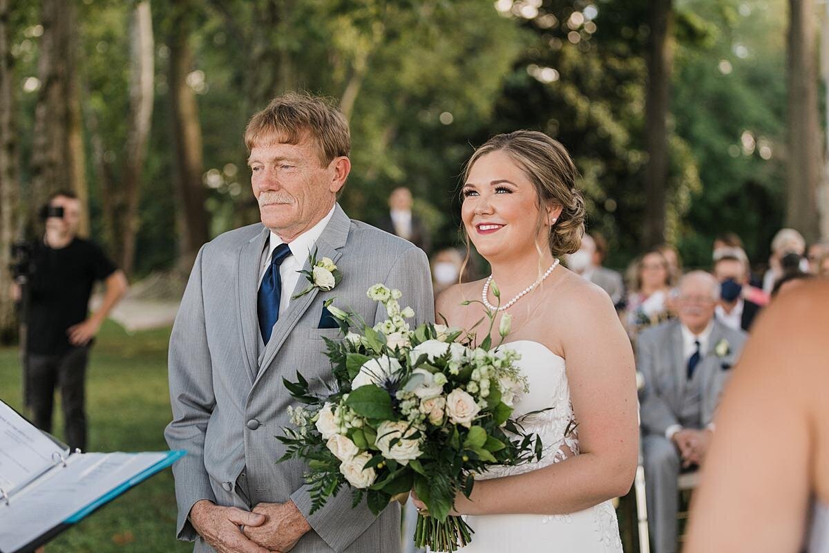 The father of the bride, dressed in a light gray suit with a white shirt and navy tie walks the bride down the aisle. The bride is wearing a strapless wedding dress with a pearl necklace and she is holding a large bouquet of white roses, white hydrangea, white peonies and greenery at The Estate at Cherokee Dock.