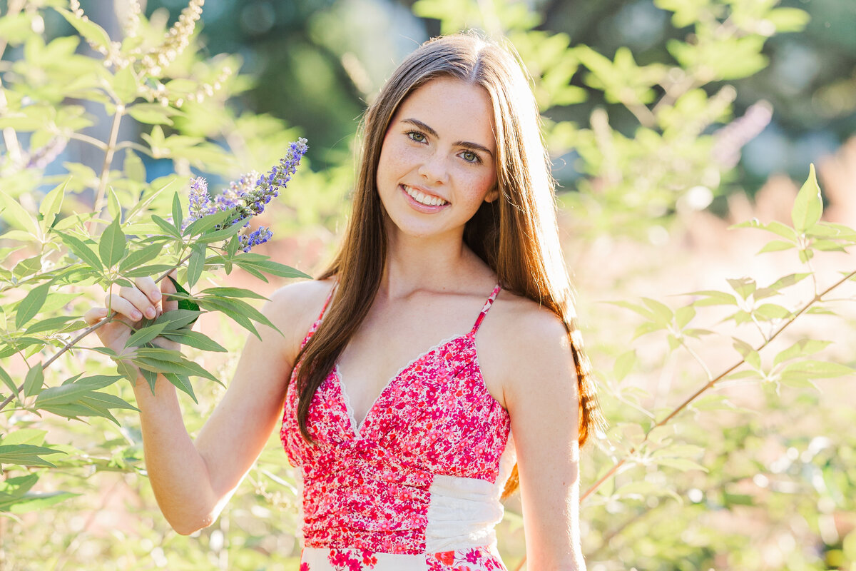 High school senior girl smiling and standing among flowering branches at sunset, wearing a pink floral dress. Captured in natural light with golden hour glow in McKinney, Texas.