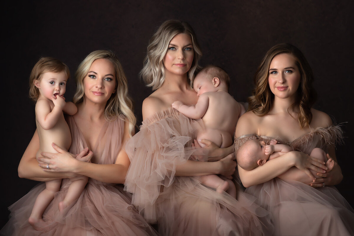 A serene family portrait by a Dallas family photographer, showing three mothers holding their babies in soft, nude-toned dresses. Each mother embraces their child, with the babies content in their arms. The backdrop features a dark, textured tone that highlights the soft elegance of the scene.