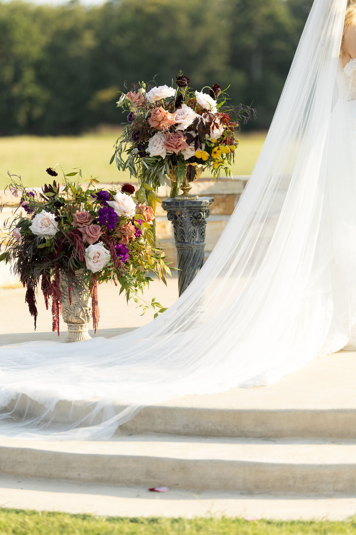 Close-up of large statement wedding ceremony urn arrangements overflowing with roses, amaranthus, greenery, and seasonal blooms in mauve, blush, burgundy, and purple, showcasing lush garden-style floral design at an outdoor ceremony altar.