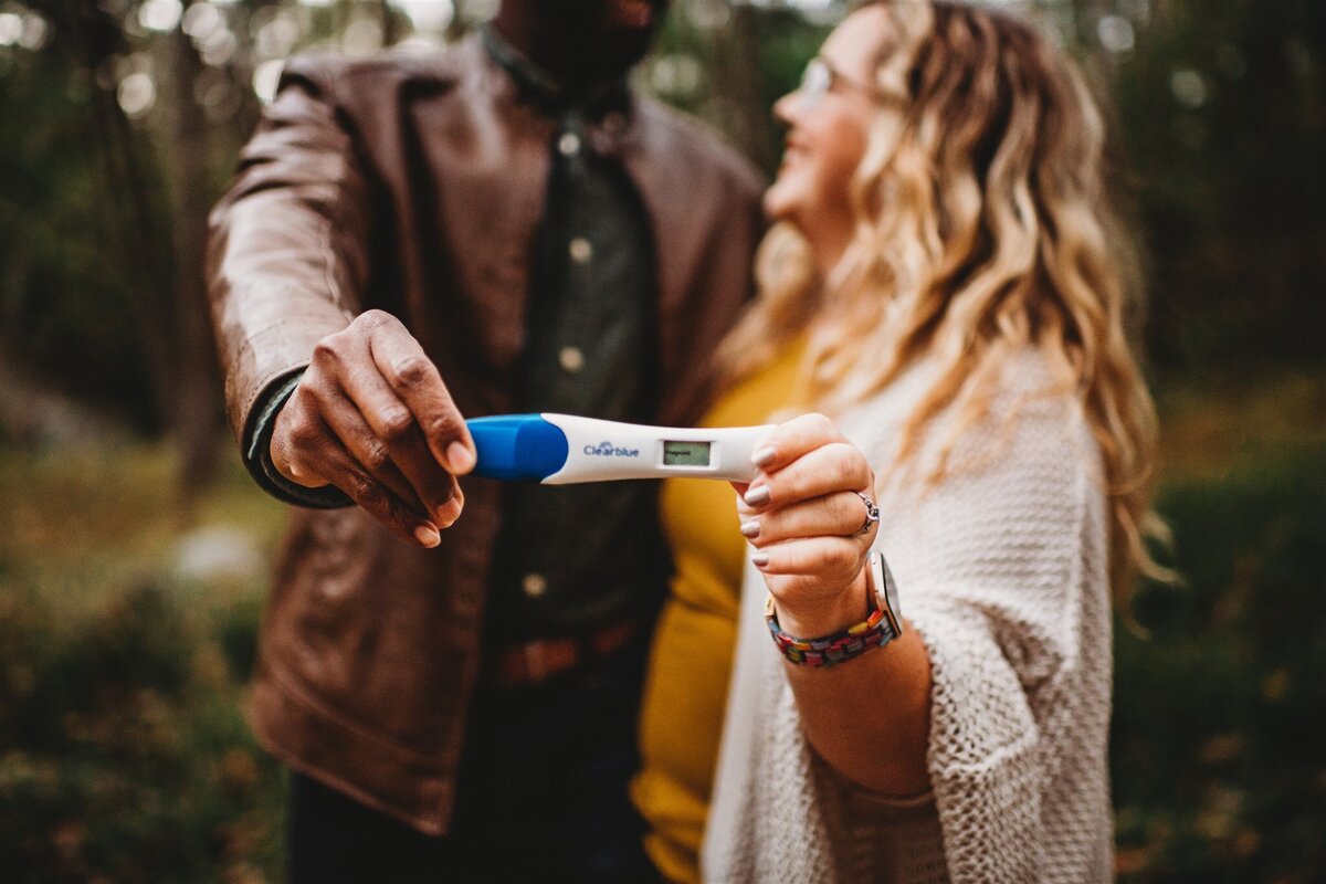 Family photographers Maryland photographs pregnancy announcement with man and woman holding a positive pregnancy test as they smile and look tog each other