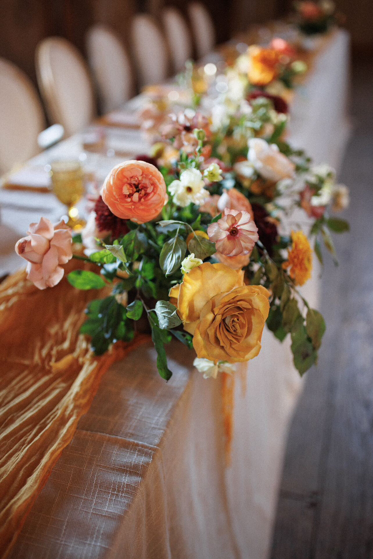 A loose centrepiece made with mustard and peach flowers sits at the edge of a long head table with a mustard yellow cheesecloth runner in the barn venue Evermore in Almonte Ontario