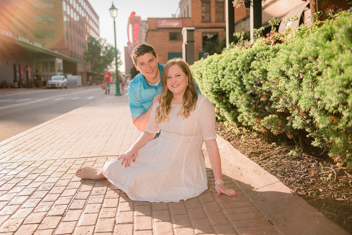Sun-filled image of engaged couple seated on brick walkway at Station Square in Pittsburgh, PA