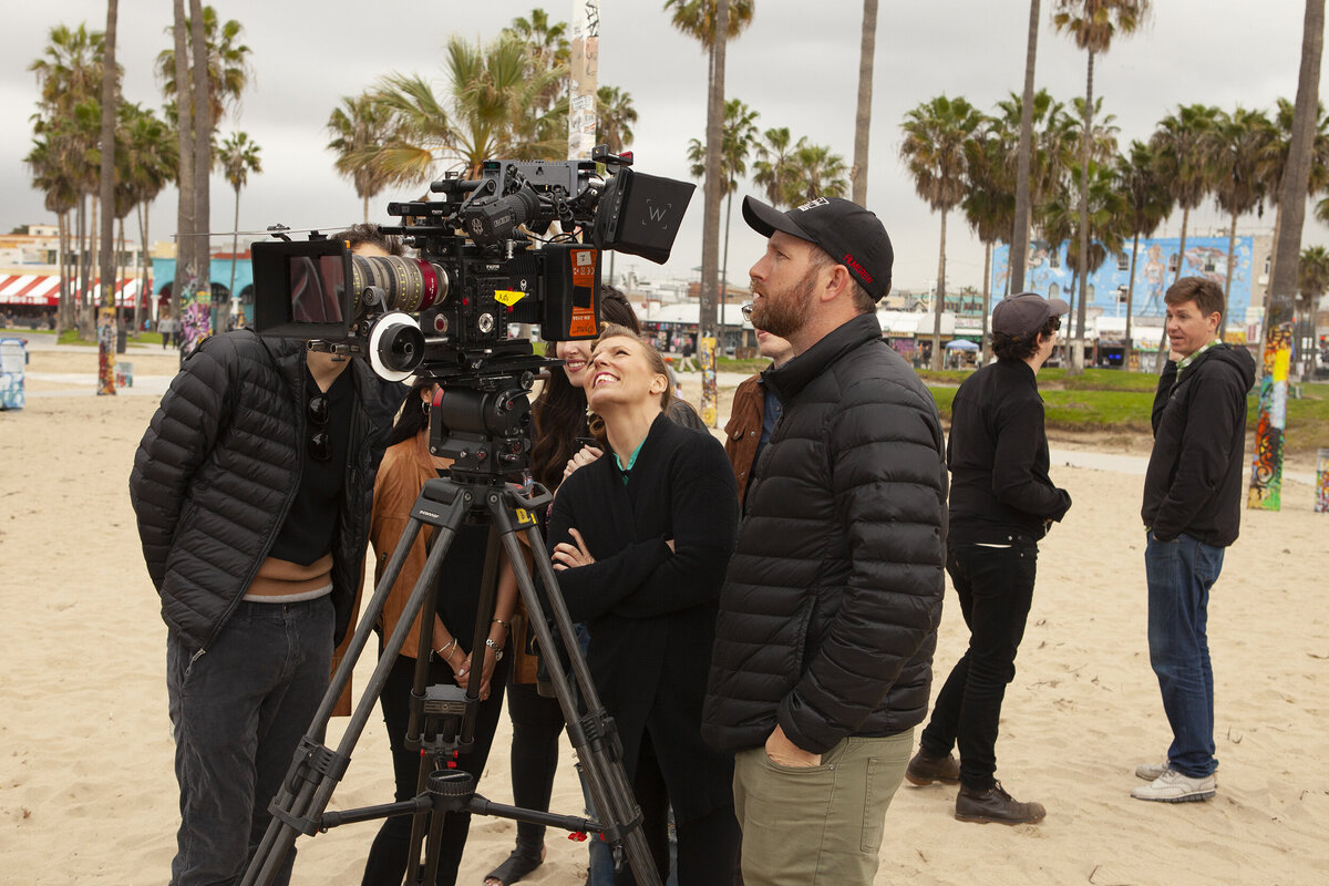 Ylva Erevall directing a TRESemmé campaign shoot on a beach, collaborating with the crew under cloudy skies and palm trees.