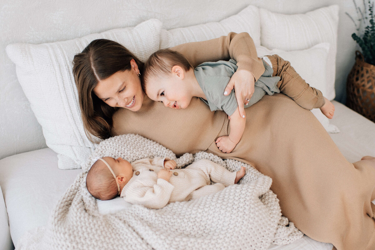 mom lying on bed looking at baby while toddler climbs over her to snuggle