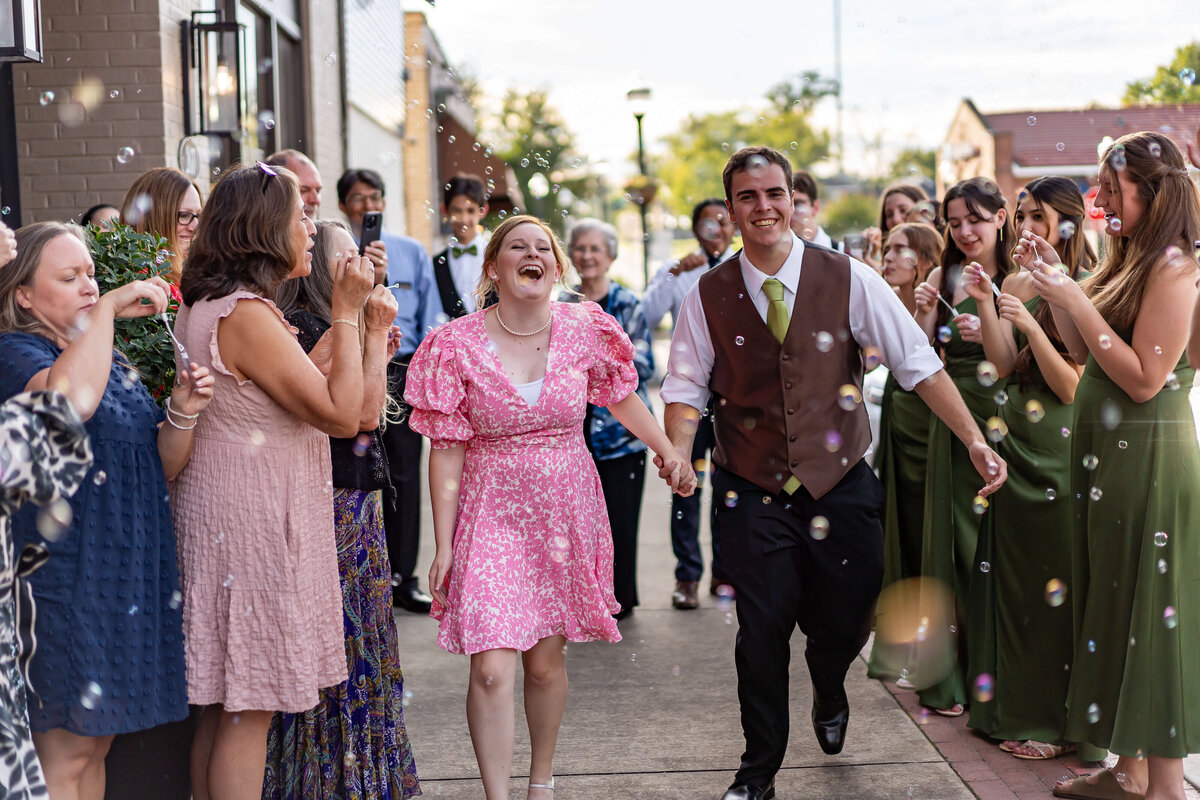 A joyful couple walks hand in hand through a crowd of smiling people blowing bubbles outdoors. The woman wears a pink dress and the man wears a vest and tie. The group celebrates, standing on either side of the couple.