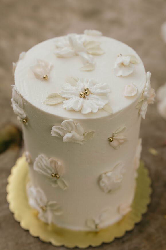 An angled shot of a white cake with piped buttercream flowers and petals in white and pale pink with pastel green leaves and gold pearl accents. 