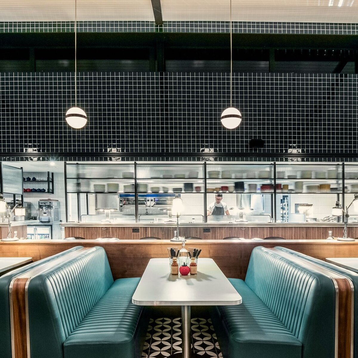 Diner Restaurant Booths with channel tufted leather benches, dining table with laminate tops and metal base. Black tiled walls and globe pendant lights in the background