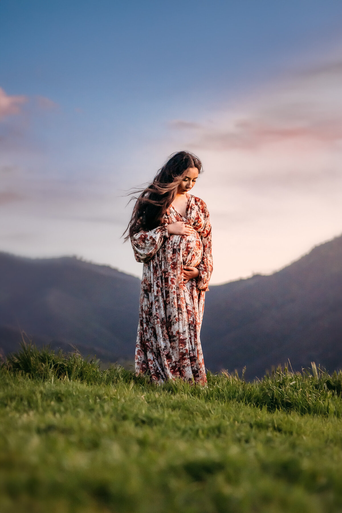 mom in floral maternity dress softly looks down at her baby bunp. the wind blows her long hair and there are mountains behind her.