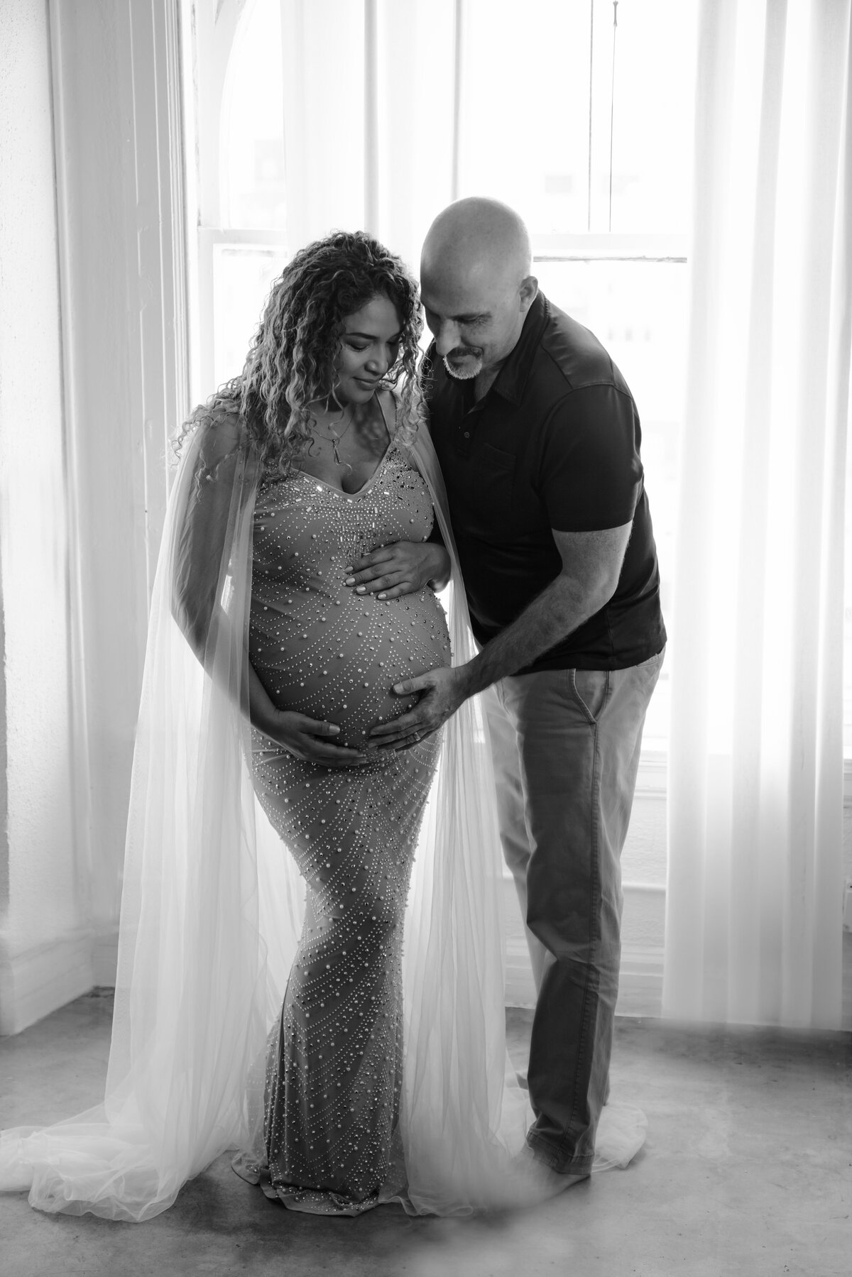 Expectant mom and dad pose for maternity photos. Mom is wearing a long beaded gown with one hand under and one hand atop of her belly. Dad is standing sideways with forehand underneath her belly. Mom and dad are looking down and smiling at her bump. Captured by Main Line photographer Liza Nicole.