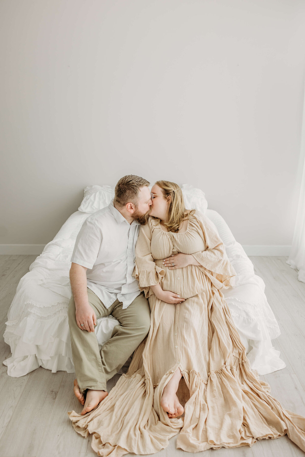Couple in beige dress and white shirt kissing on bed expecting baby near Eau Claire, WI