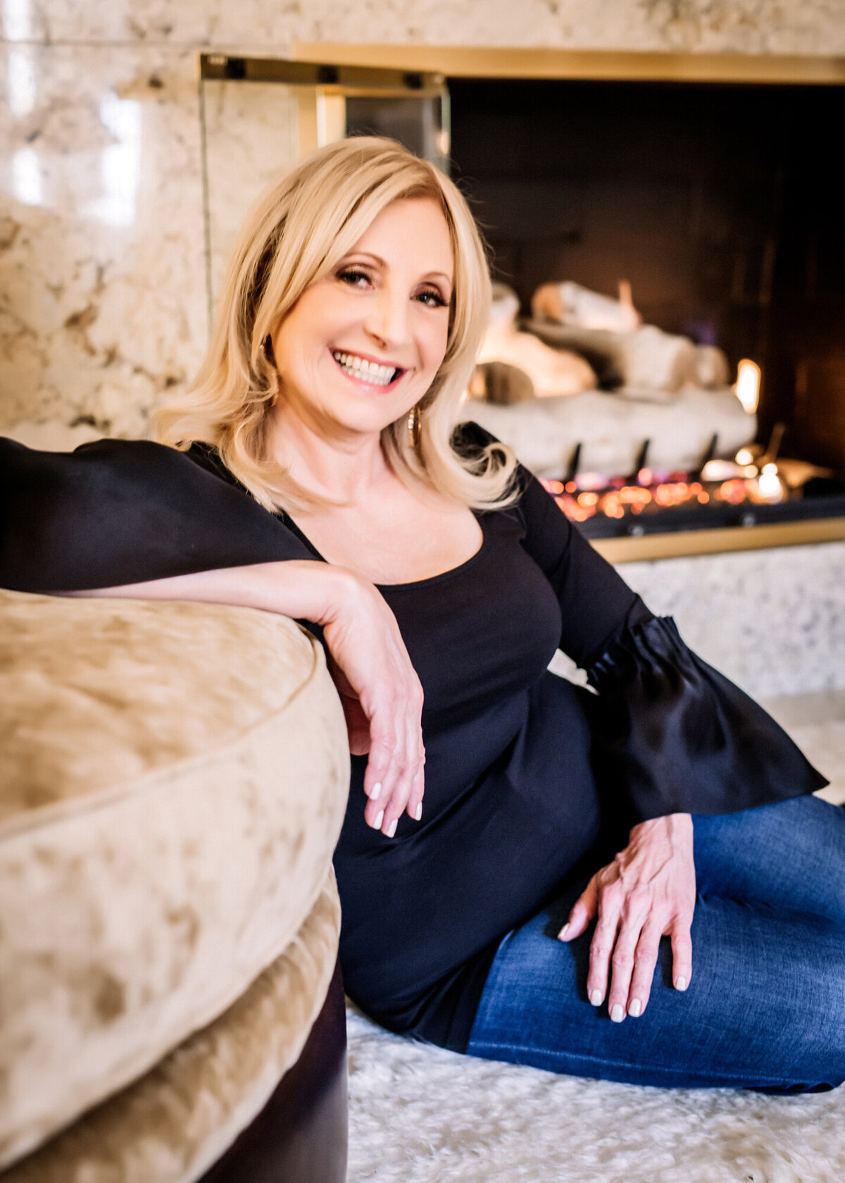 A female dating portrait client. sitting on the floor, leaning against a beige chair in front of her fireplace, smiling at the cameras