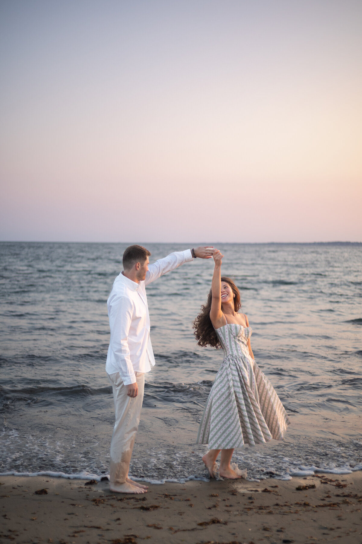Collins Beach Newport RI | Kelsey Sheehan Photography Timeless Rhode Island Weddings | A couple dances joyfully on a beach at sunset. The man in a white shirt twirls the woman in a flowy dress. The ocean's waves gently touch the shore.