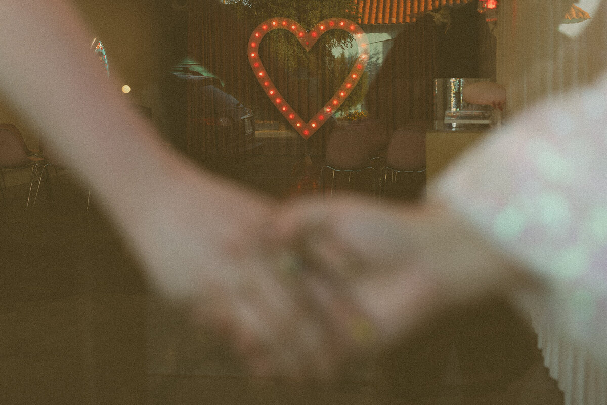Close-up of a couple's clasped hands with a neon heart sign glowing in the background