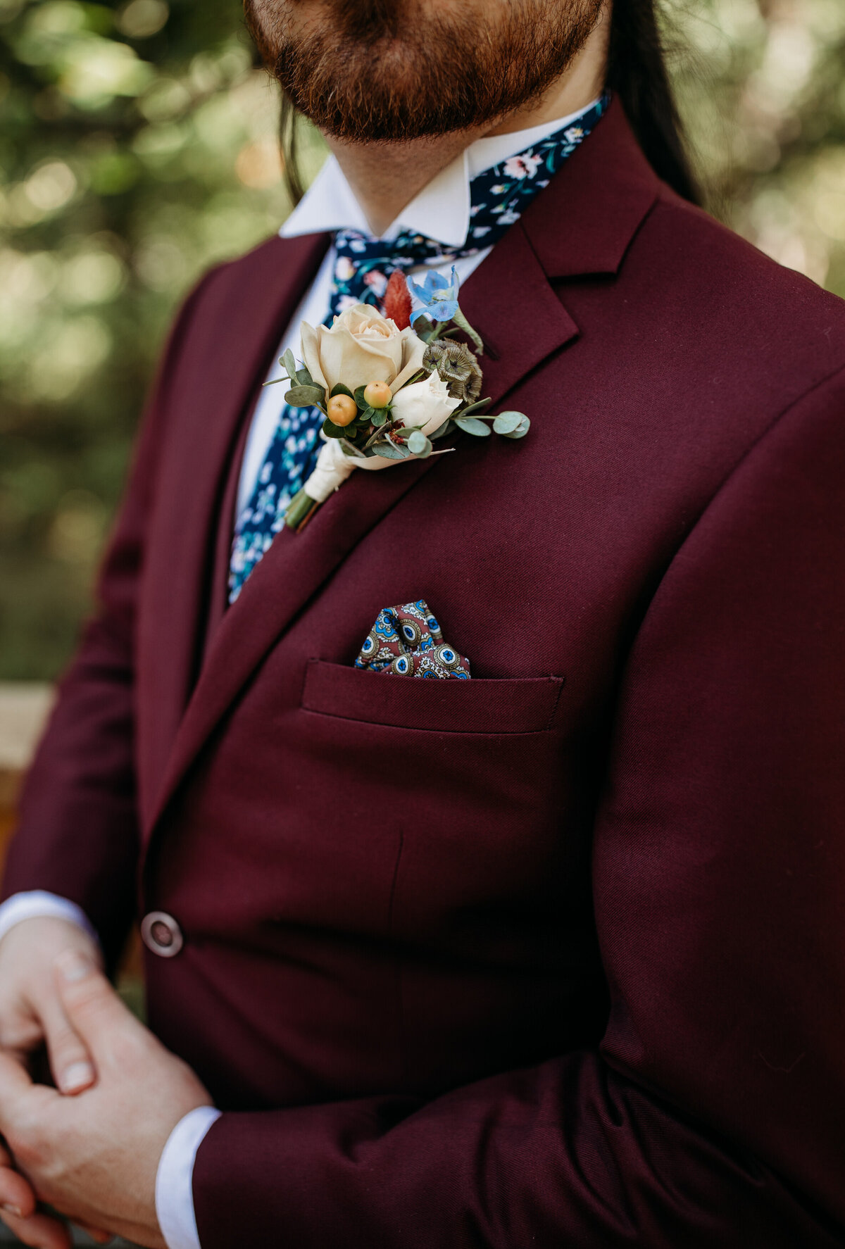 Close-up of a maroon suit jacket with a vibrant floral tie and a boutonniere featuring a rose and greenery
