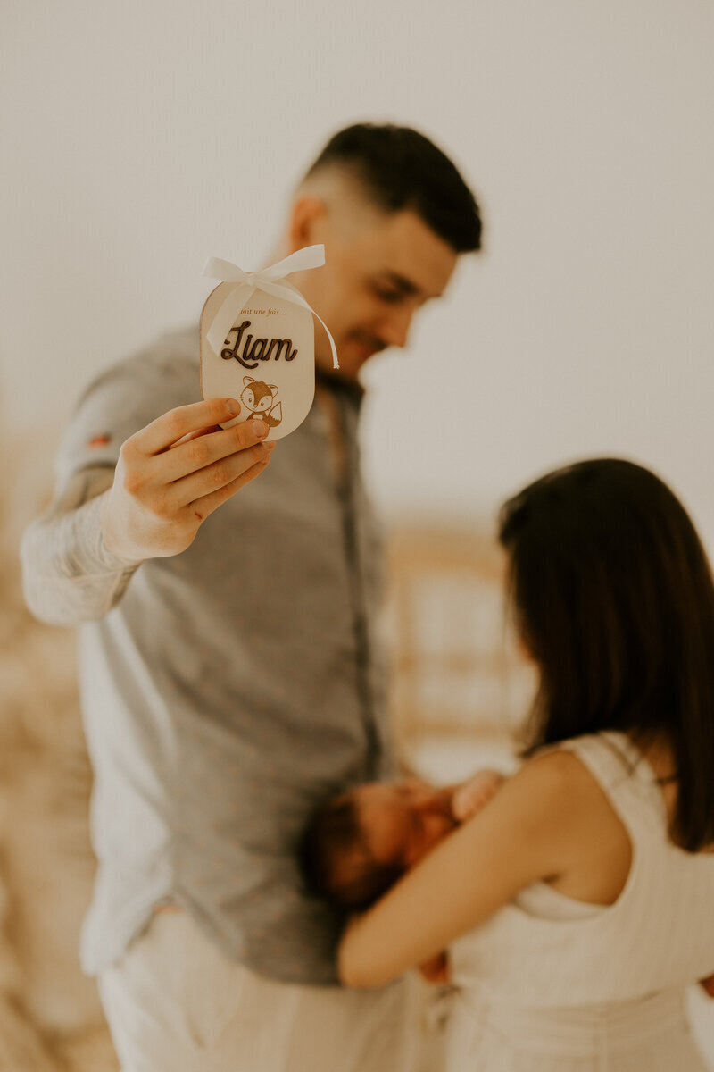 Maman portant bébé dans ses bras pendant que monsieur montre au premier plan une décoration en bois avec le prénom de bébé pour un shooting photo naissance.