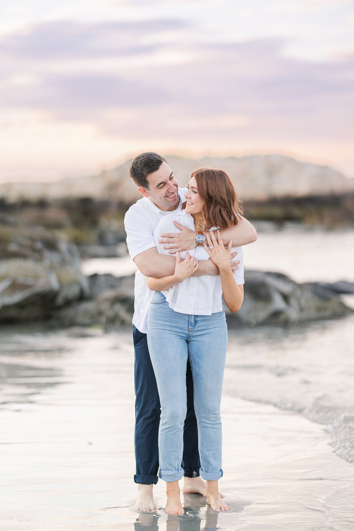 A man hugging a woman from behind and laughing during their sunset Wingaersheek Beach engagement pictures.
