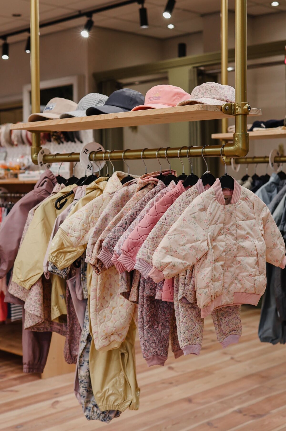 Kinderkleding hangt aan een gouden rek, petten op een plank erboven, in een winkel met houten vloer.