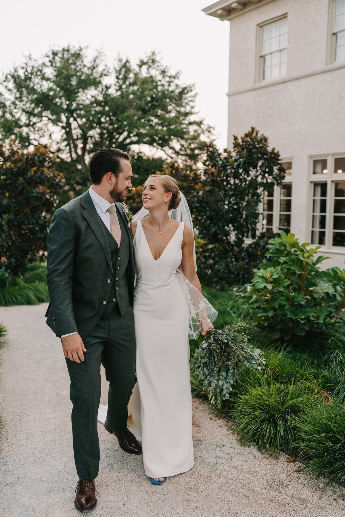 A photograph in color of Colby and Jack on their wedding day at Commodore Perry Estate in Austin, Texas. The photograph shows the couple walking arm in arm while they look at each other, smiling. The bride is wearing a classic white satin wedding gown, blue high heeled shoes and a fingertip length veil with her light hair pulled back in a bun. The groom is wearing a dark gray three piece suit with a blush colored tie. There is a tall oak tree in the distance, two small magnolia trees and other green shrubs on either side of the gravel path they are walking on. On the right, is the side of a historical mansion with off-white stucco and tall windows with square panes. Wedding photography by Stacie McChesney/Vitae Weddings.
