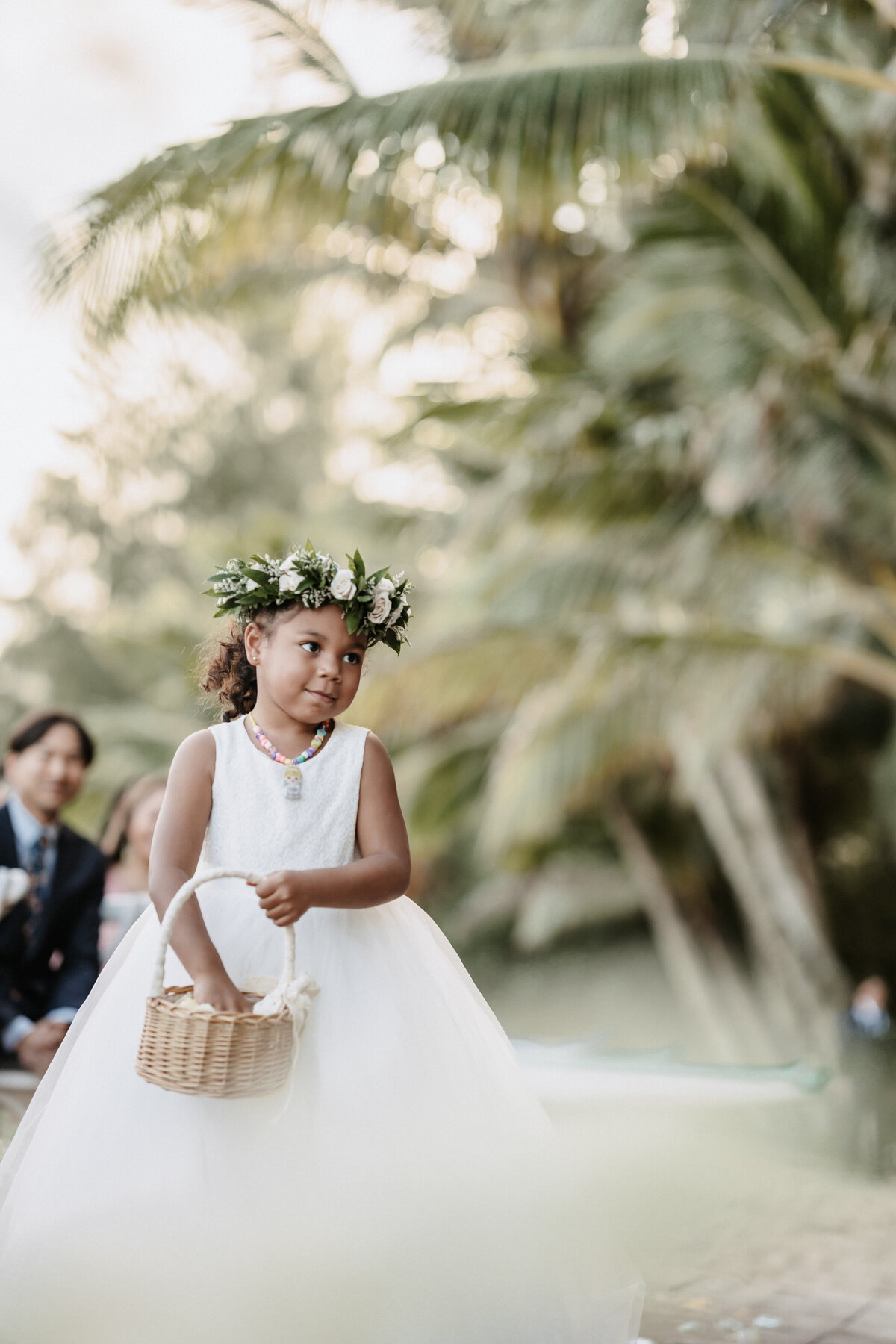 Flower girl walking down the aisle in ivory dress with hand in basket on the beach and palm trees behind her