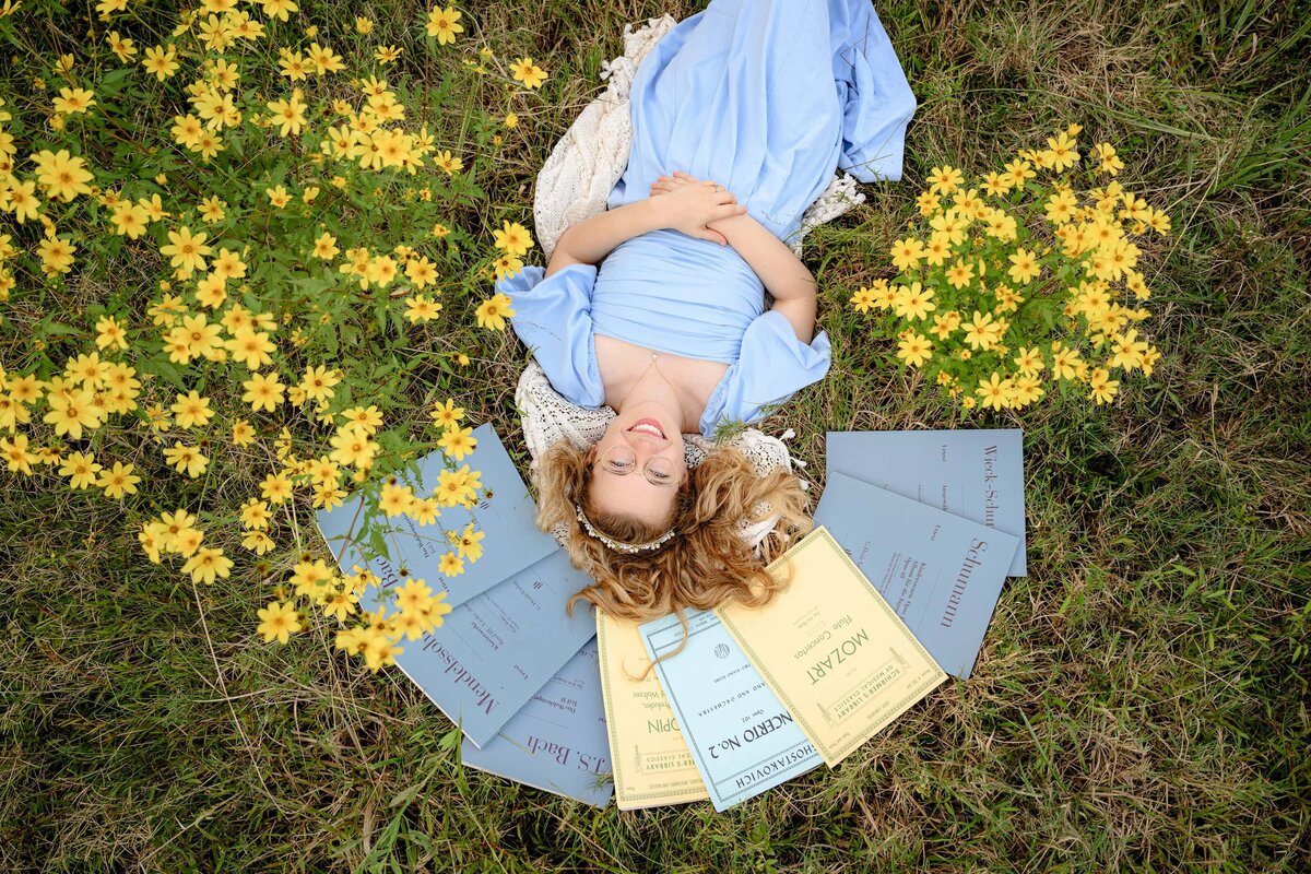 Charlotte NC senior in high school lays in a field with yellow flowers wearing a blue dress with her musical scores around her