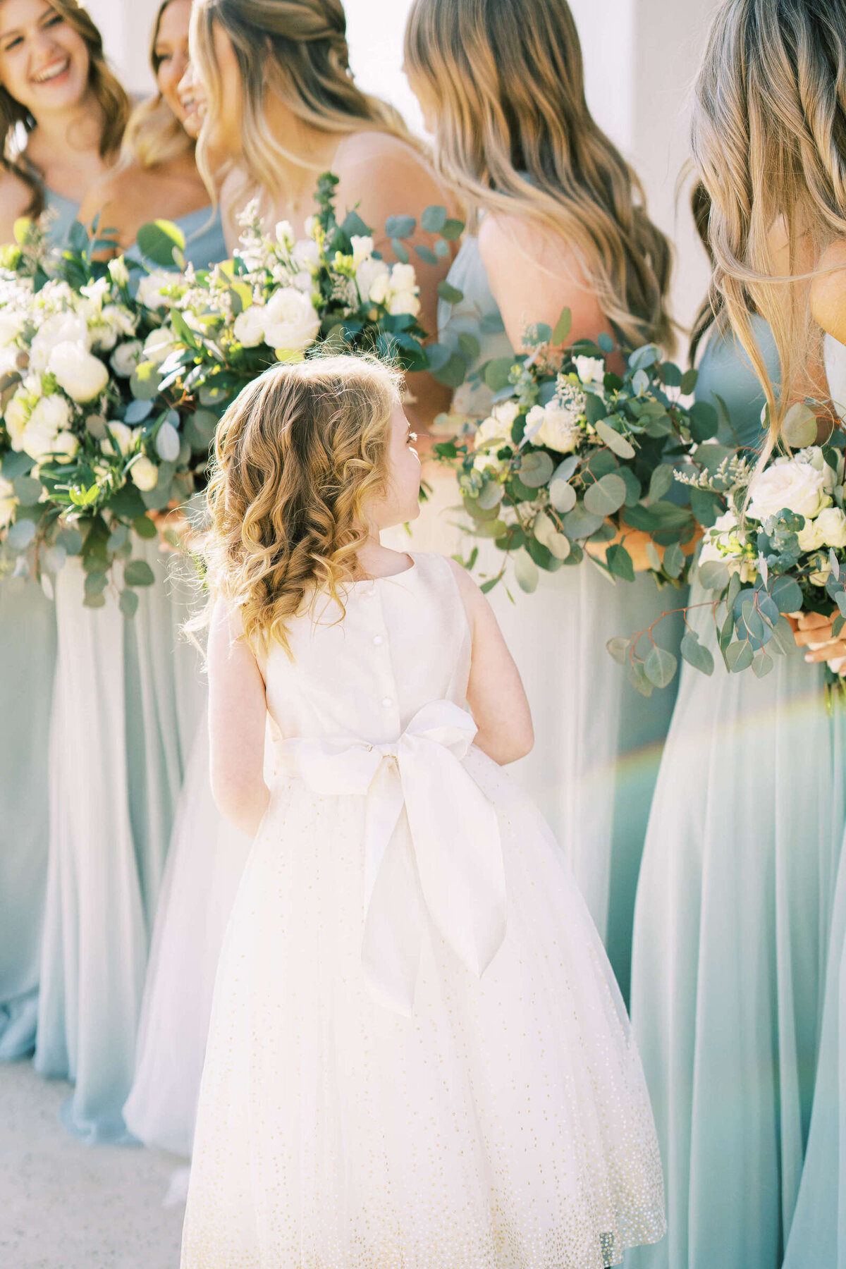 Curly haired flower girl in white flowy dress stands next to bridesmaids