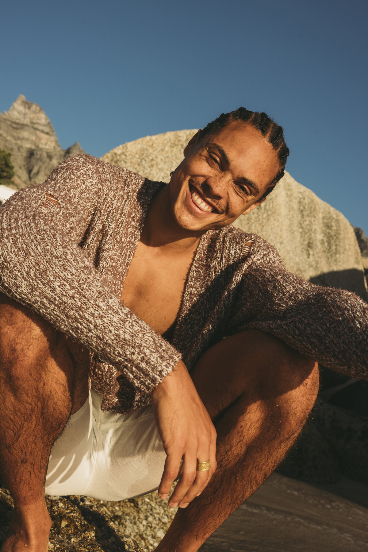 Portrait of a smiling male model with braids wearing a knitted brown sweater and white shorts, seated on rocks during golden hour in Cape Town.