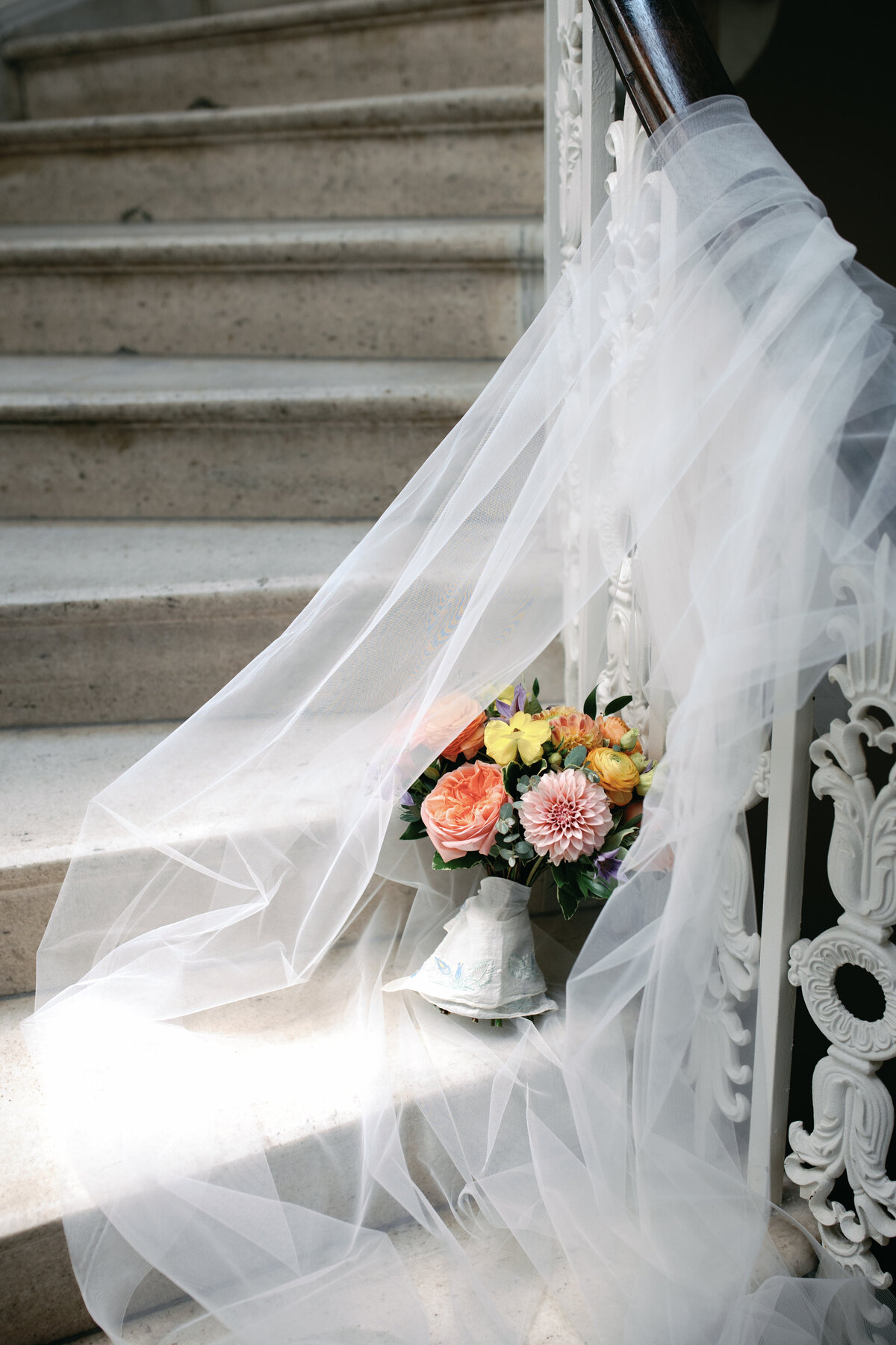 Vibrant bridal bouquet with pink, orange, and ivory flowers placed on marble staircase beneath flowing cathedral veil.