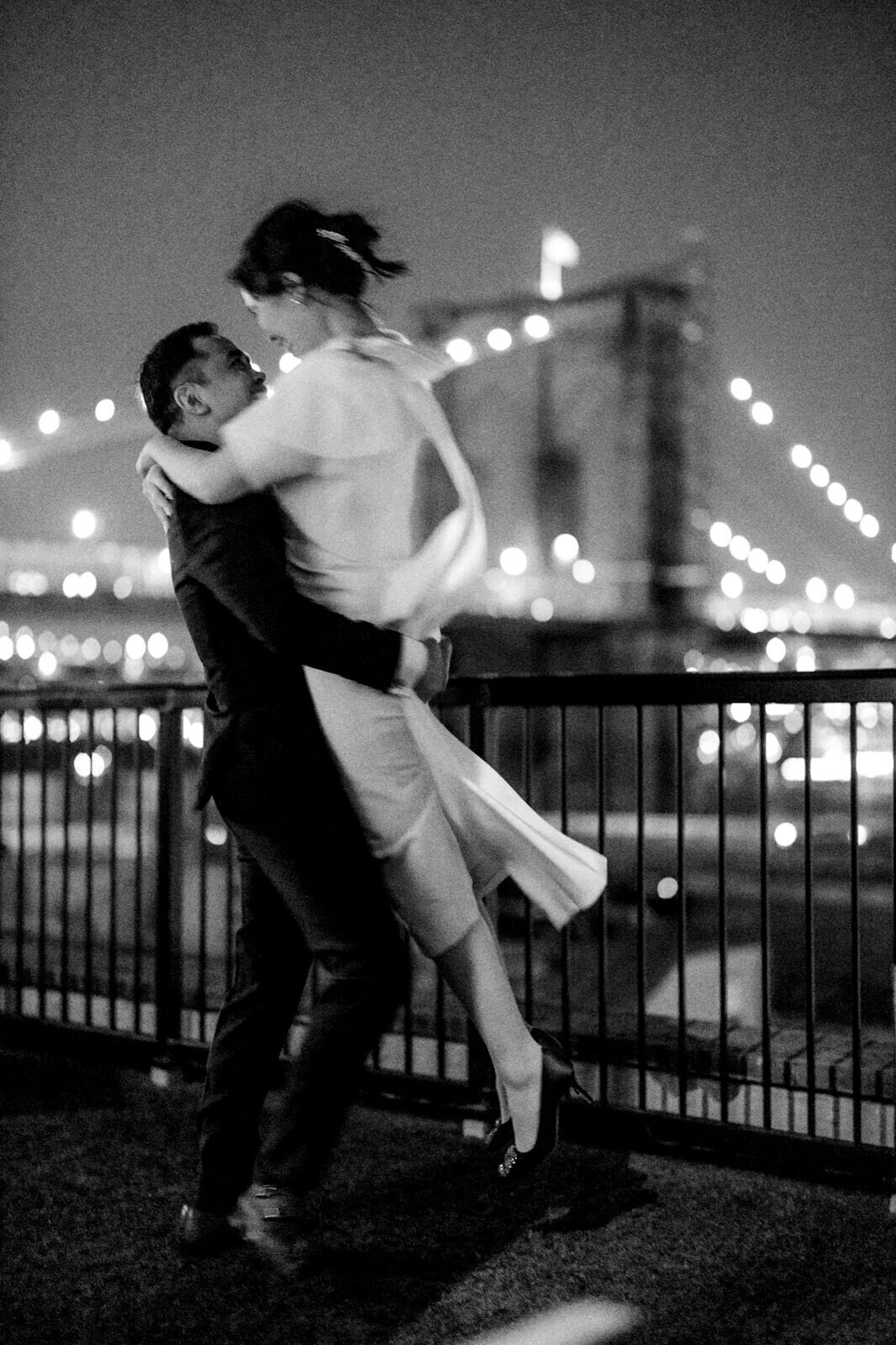 A night view of the groom carrying the bride on a veranda overlooking the Brooklyn Bridge.