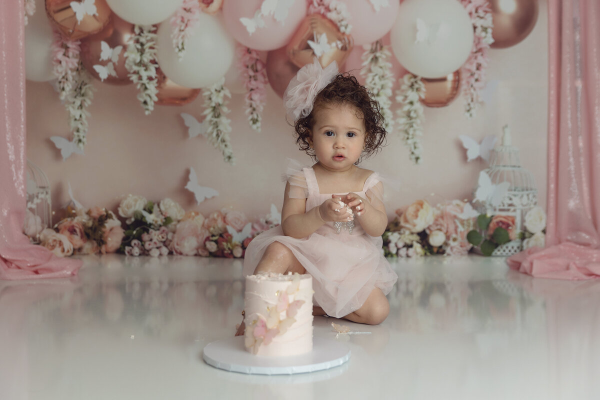 A toddler girl in a pink dress explores a pink cake during her first birthday