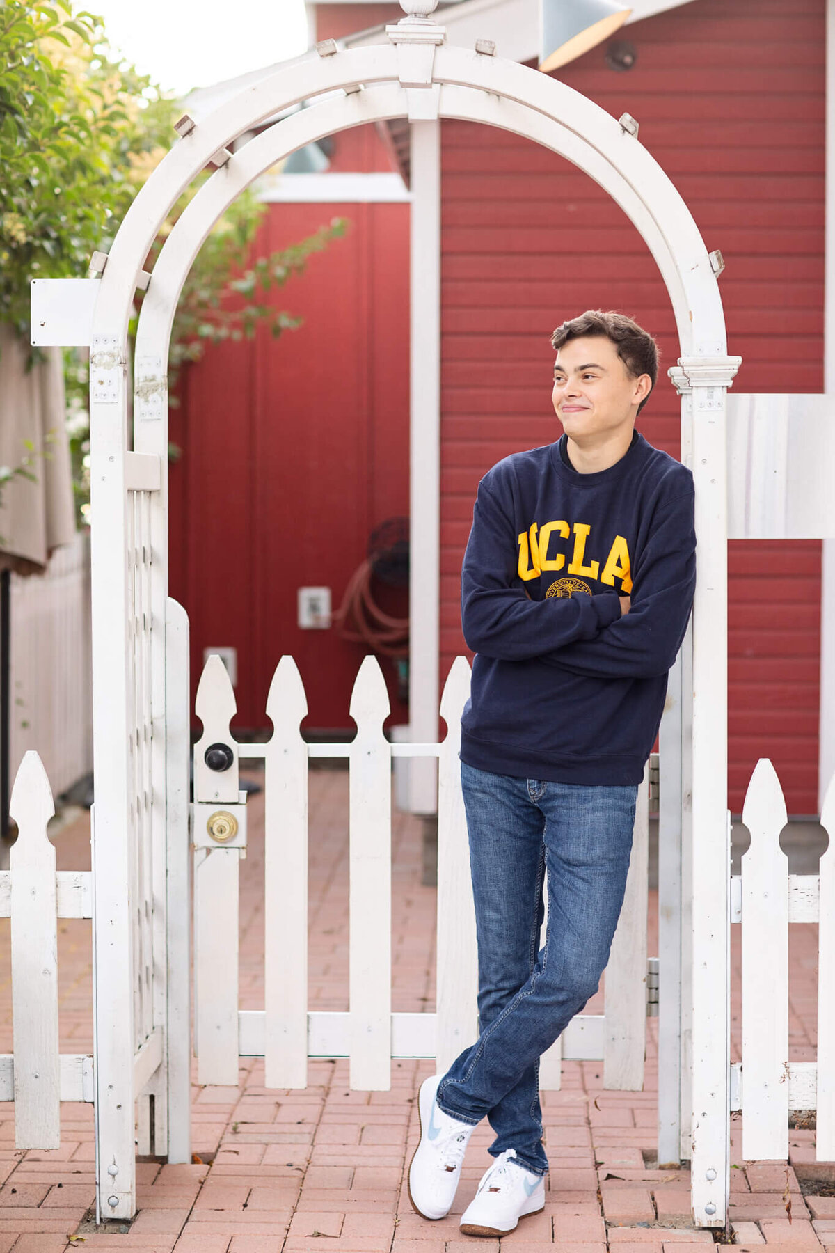 A person wearing a UCLA sweatshirt stands casually with arms crossed under a white archway. They are in front of a red building, next to a white picket fence. The person looks to the side with a slight smile.