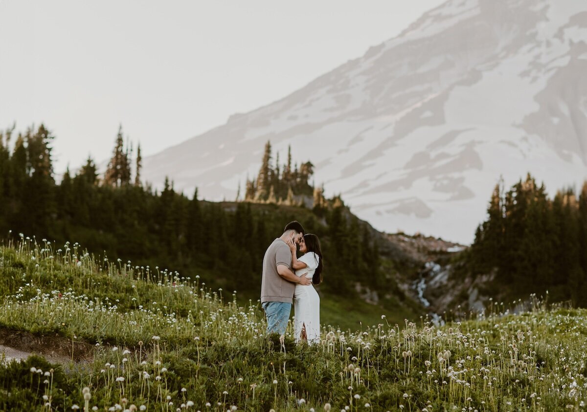 A man in jeans and a shirt holds his fiancee close in a flower meadow in her white dress