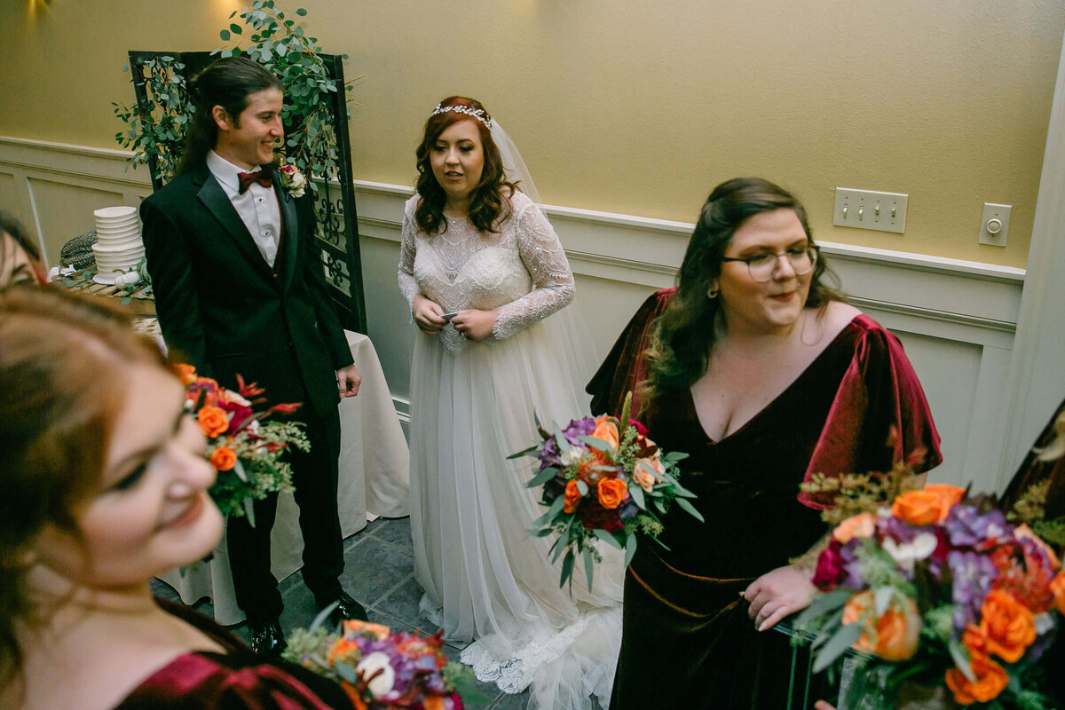 A bride in a white dress stands next to a groom in a black suit, surrounded by bridesmaids in burgundy dresses holding colorful bouquets at an indoor room inside a wedding venue.