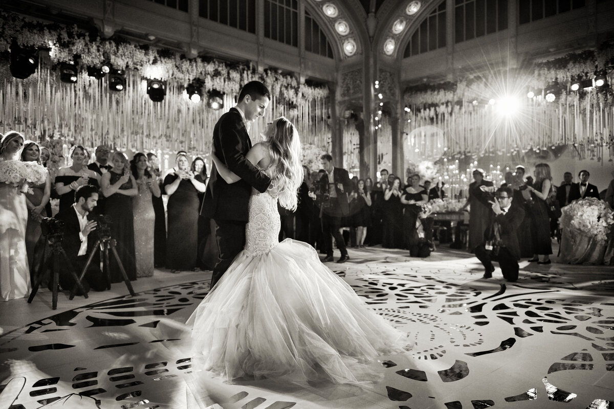 The photo captures a couple sharing their first dance at their wedding in an elegant, grand hall at The New York Public Library in New York City. The bride looks stunning in a flowing gown, while the groom is dressed in a sharp suit. Guests can be seen cheering them on, with the venue beautifully decorated and the lights shining, creating a celebratory and romantic atmosphere. The videographers from East West Films were on hand to document this special moment. The photograph was taken by photographer Christian Oth.