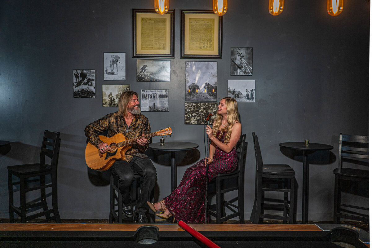 Two people sitting in high top chairs singing and playing guitar together 