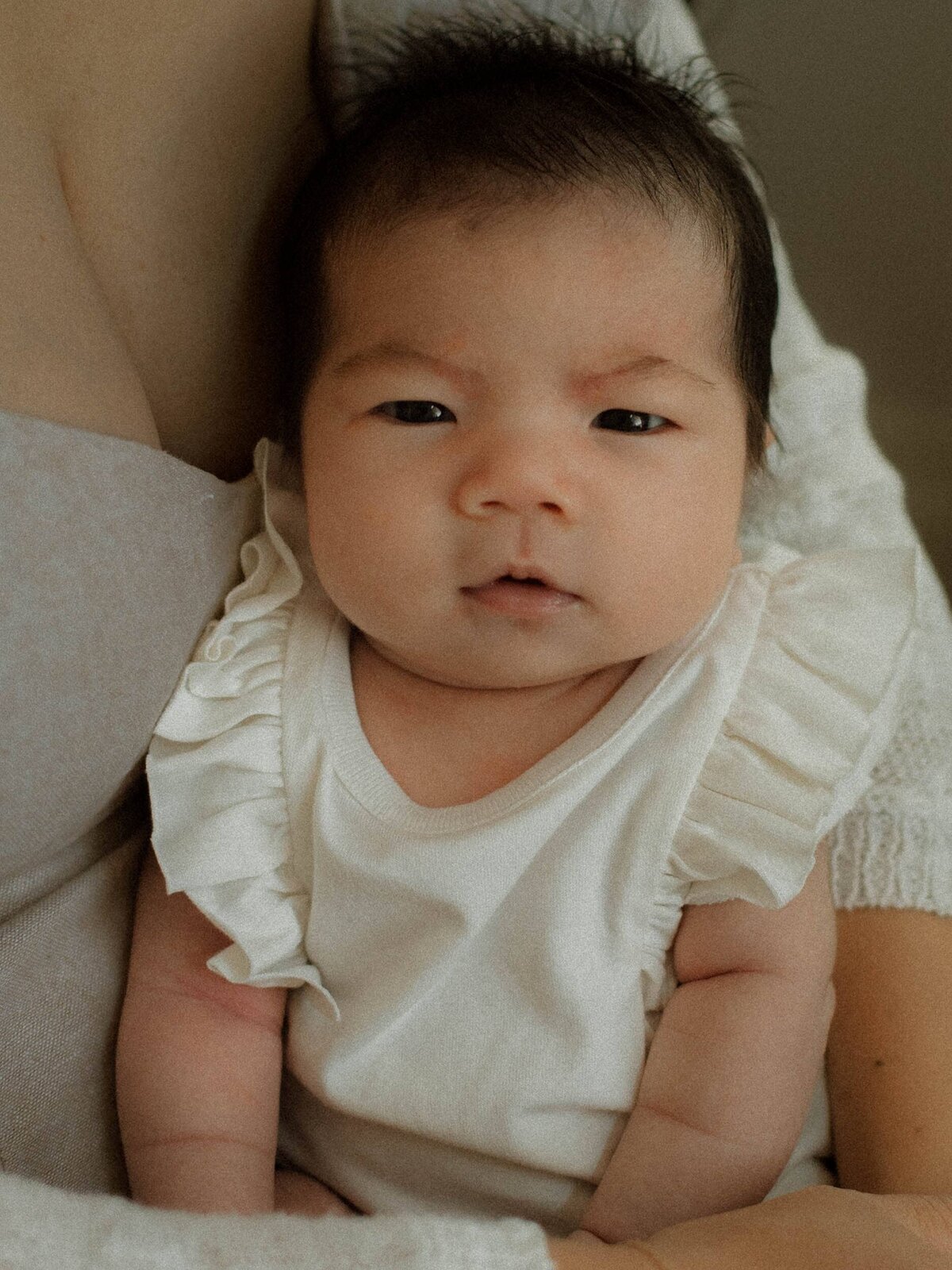 Baby portrait looking directly at the camera in white romper while sitting on mom's lap.