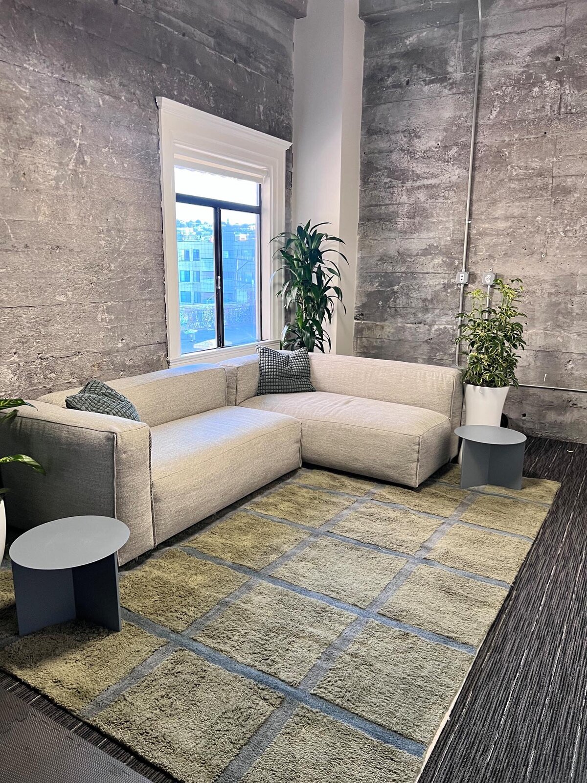 Cozy corner lounge with a light gray sectional sofa, grid-patterned rug, indoor plants, and exposed concrete walls.