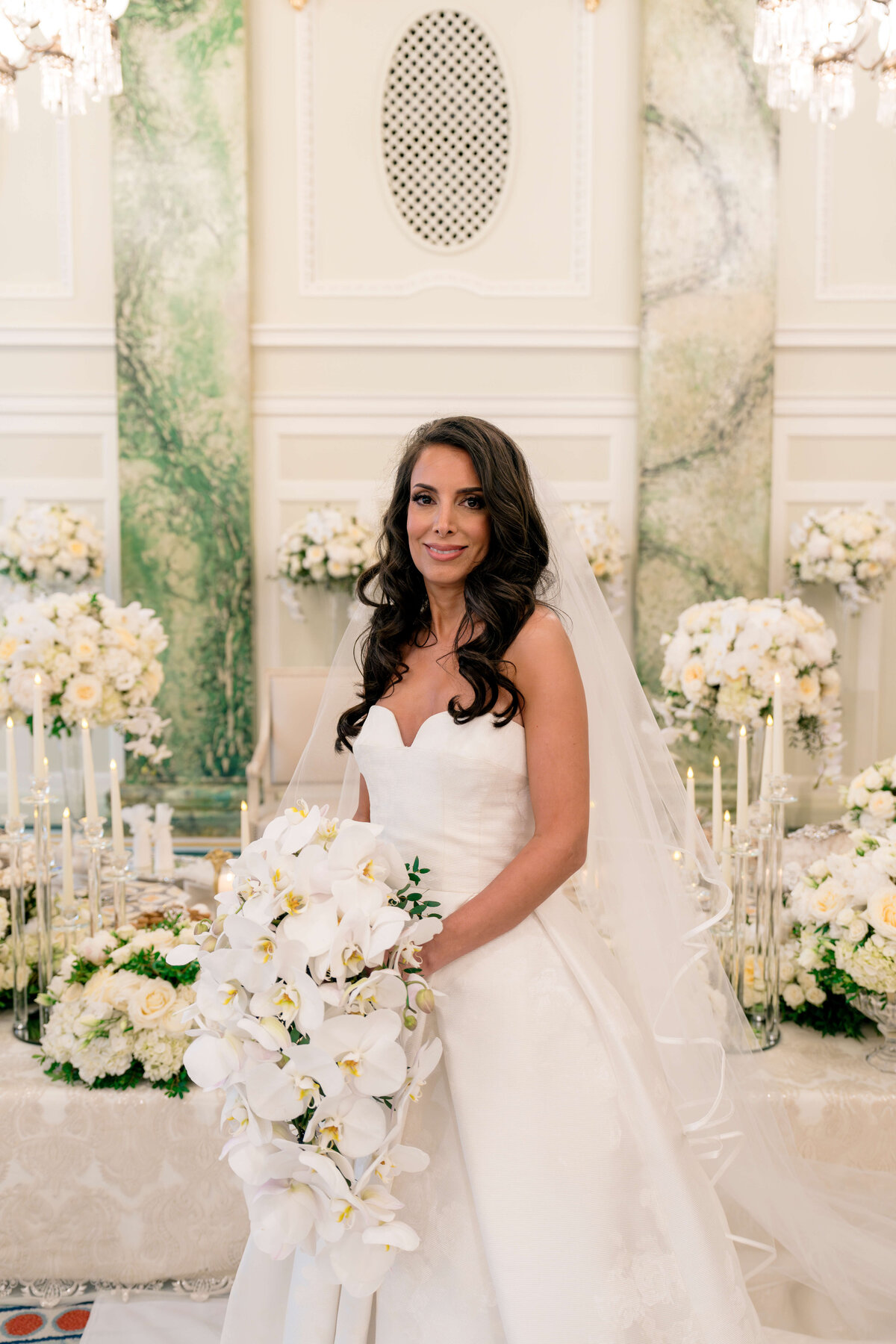 A bride in a strapless white wedding gown holds a cascading bouquet of white orchids. She is smiling and stands inside a decorated venue with white flowers and soft green accents. Chandelier lights hang above, adding elegance to the setting.