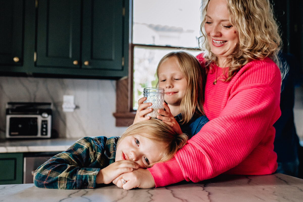 A mother and two children in the kitchen, with one child leaning on the countertop while the other holds a glass jar. The mother, smiling, gently embraces them.