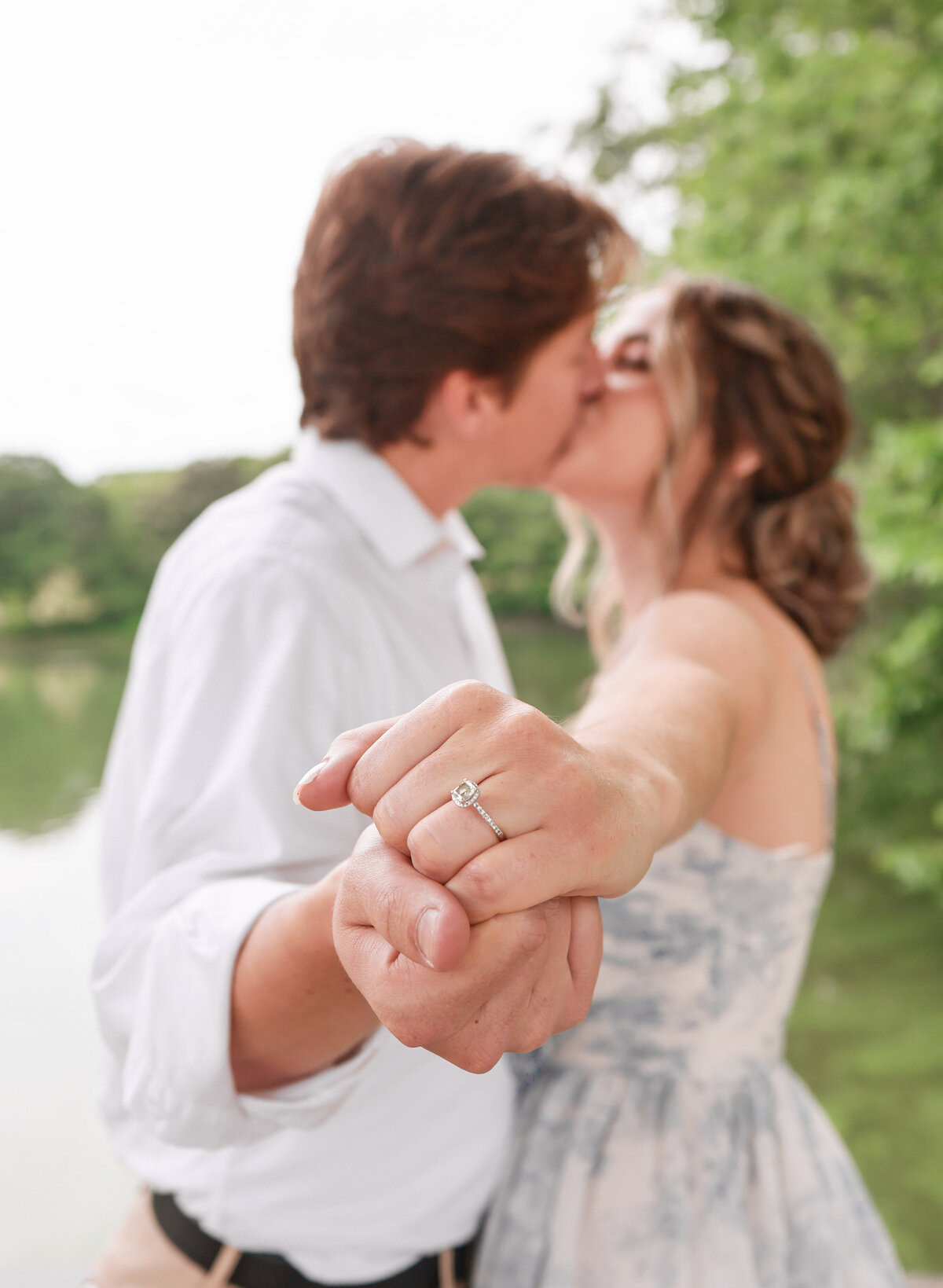 man wearing white shirt and woman wearing blue and white floral dress kissing and holding hands out showing off diamond engagement ring